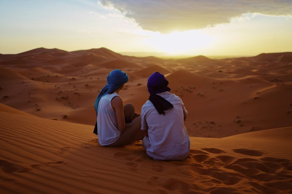 camel trekking refreshments Tourists sit in the serene Sahara desert, immersed in the breathtaking colors of a sunset sky after their memorable Merzouga Camel Trek adventure.