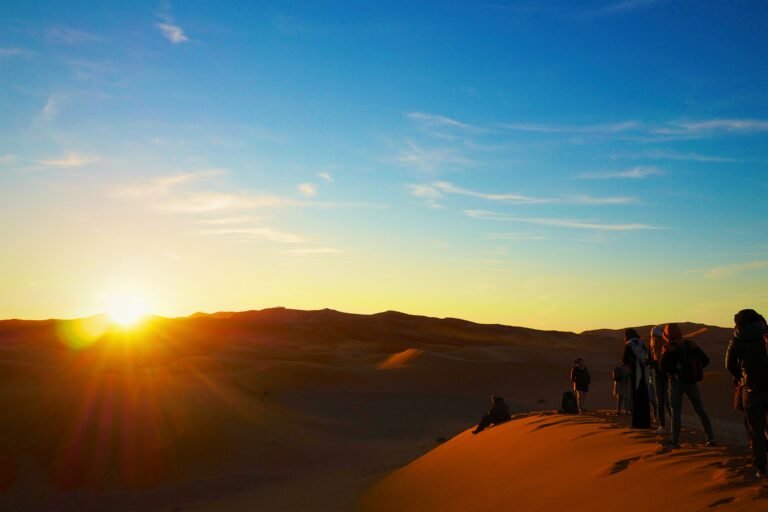 A line of camels carrying travelers walks across the sand dunes at sunrise, marking the start of an unforgettable Dades Gorge Trip through the Moroccan Sahara.