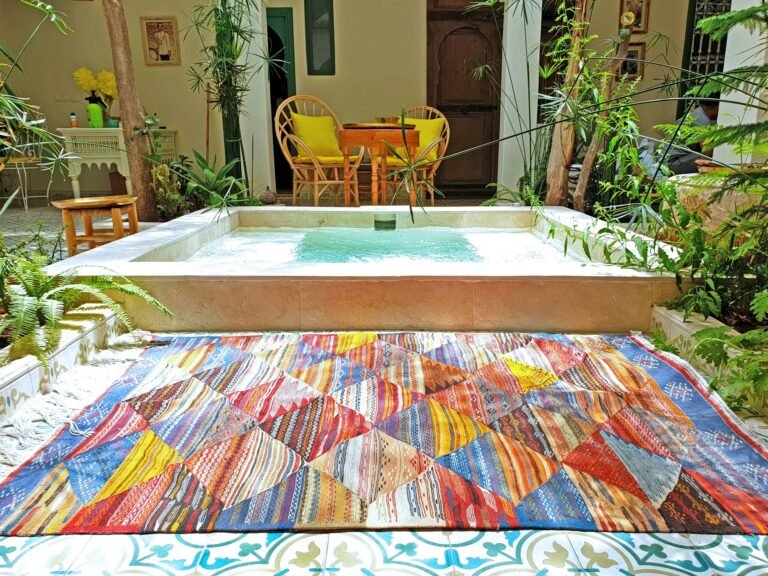 The image shows a tranquil Moroccan home courtyard, where a small, ornately framed water fountain takes center stage. Warm sunlight shimmers on the calm water's surface, surrounded by traditional mosaic tiling. The rustic beauty of the scene is completed by a luxurious traditional Moroccan rug elegantly draped beside the fountain, adding warmth and rich color to the space. The rug forms the focal point that connects the natural and architectural elements, creating a vista that evokes comfort and authentic beauty. This simple yet powerful arrangement is a perfect example of how to integrate Moroccan Rugs into exterior design to create an atmosphere of relaxation and rustic elegance.