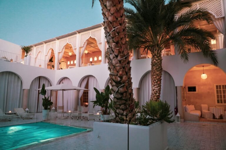 This breathtaking image captures the essence of a Luxury Riad Marrakech, showcasing its traditional architecture with ornate arches and detailed zellij tilework. A serene turquoise pool reflects the lush greenery and the warm glow of lanterns, creating a perfect oasis of tranquility and authentic Moroccan elegance in the heart of the city.