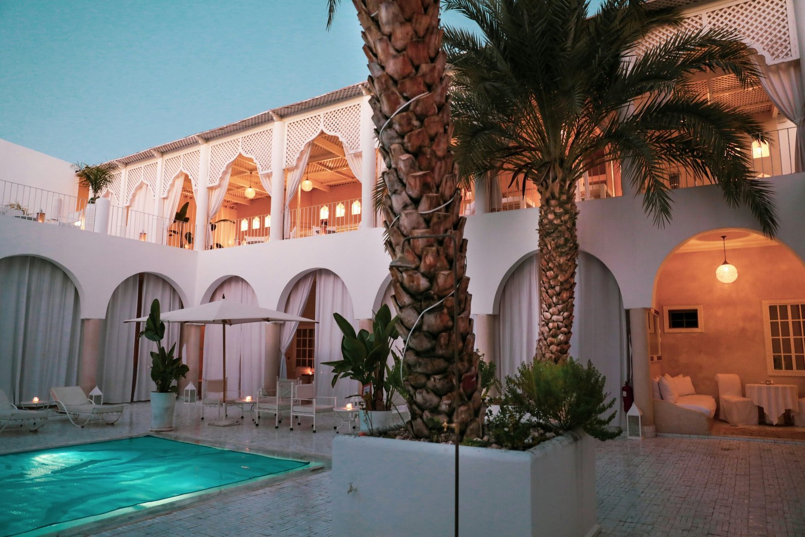 This breathtaking image captures the essence of a Luxury Riad Marrakech, showcasing its traditional architecture with ornate arches and detailed zellij tilework. A serene turquoise pool reflects the lush greenery and the warm glow of lanterns, creating a perfect oasis of tranquility and authentic Moroccan elegance in the heart of the city.