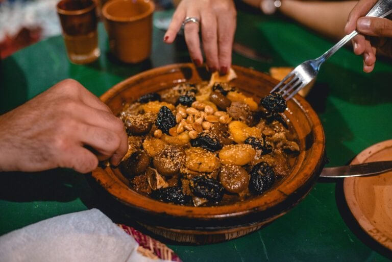 At the heart of bustling Marrakech, this image captures the very essence of the sensory experience offered by a Food Tour Marrakech. The focus is on a warm, traditional tagine, overflowing with tender lamb and sweet Fassi prunes, with an aromatic steam that suggests it has just been slow-cooked to perfection. Hands instinctively reach toward the dish in a spontaneous gesture of sharing and delight, ready to take that first delicious bite. This intimate moment, where the boundaries between visitor and authentic cuisine dissolve, is precisely what a Food Tour Marrakech provides: a profound sense of connection and enjoyment of Morocco's true flavors.